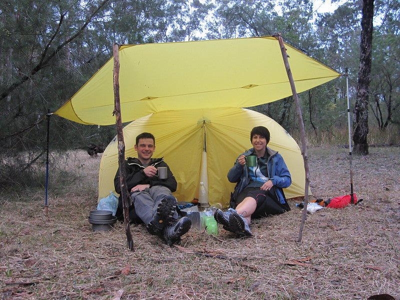 Two campers enjoying drinks under a yellow tent in the woods.